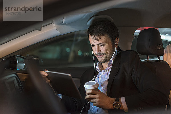 Geschäftsmann sitzt nachts im Auto  benutzt digitales Tablet  trinkt Kaffee