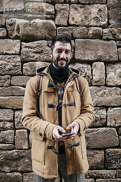 Spanien  Igualada  Porträt eines lächelnden Mannes  der mit einem Mobiltelefon an einer Steinmauer steht
