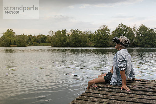 Älterer Mann  der mit einer VR-Brille auf einem Steg an einem See sitzt