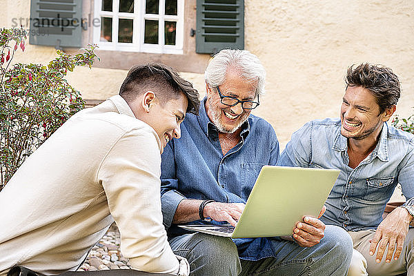 Drei glückliche Männer unterschiedlichen Alters mit Laptop im Garten