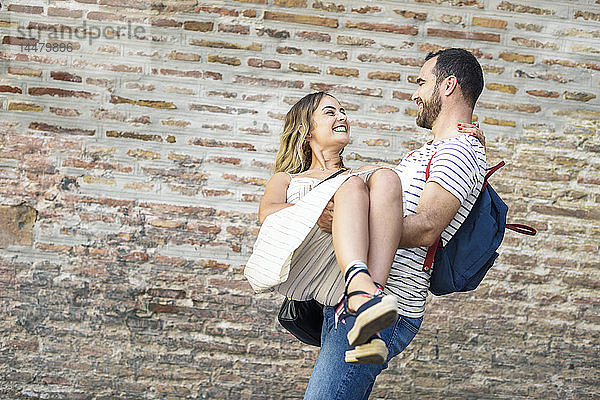 Glücklicher Mann mit Rucksack trägt Freundin an Ziegelmauer