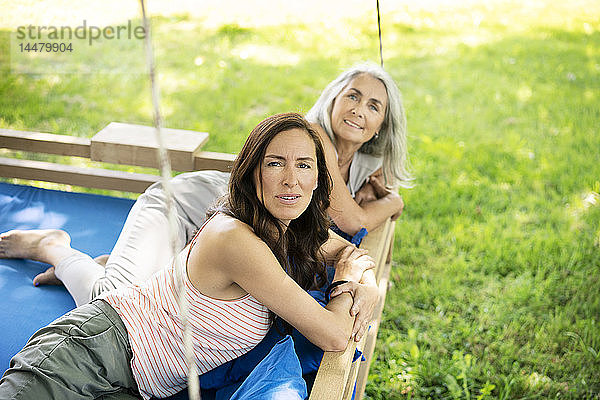 Porträt von zwei lächelnden Frauen  die sich auf einem hängenden Bett im Garten entspannen