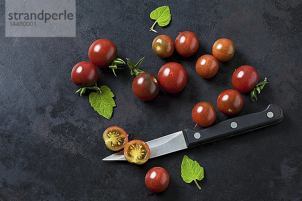Ganze und in Scheiben geschnittene rispische Tomaten 'Black Cherry'  Blätter und Küchenmesser auf dunklem Grund