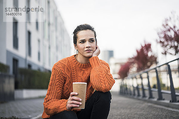 Porträt einer nachdenklichen Frau mit Kaffee zum Mitnehmen in orangem Strickpullover im Freien