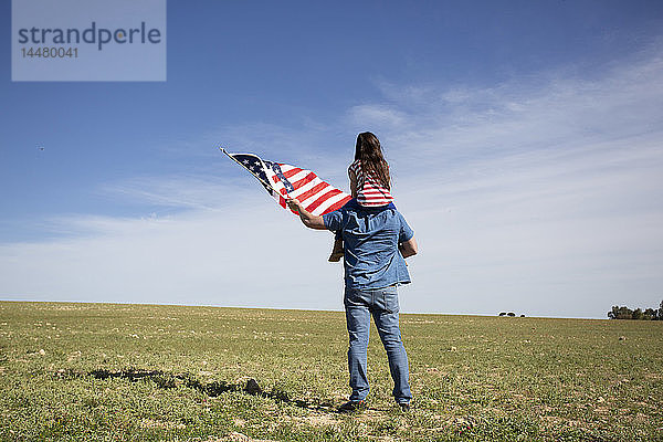 Mann mit Tochter und amerikanischer Flagge auf Feld in entlegener Landschaft stehend