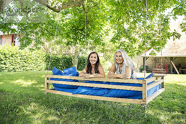 Porträt von zwei lächelnden Frauen  die sich auf einem hängenden Bett im Garten entspannen