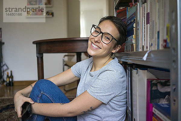 Glückliche junge Frau sitzt zu Hause und lehnt auf einem Bücherregal