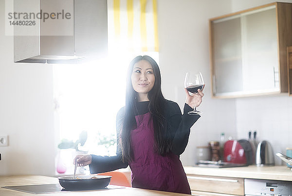Junge Frau kocht zu Hause und hält ein Glas Rotwein