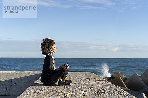 Frau sitzt auf Mauer vor dem Meer und meditiert