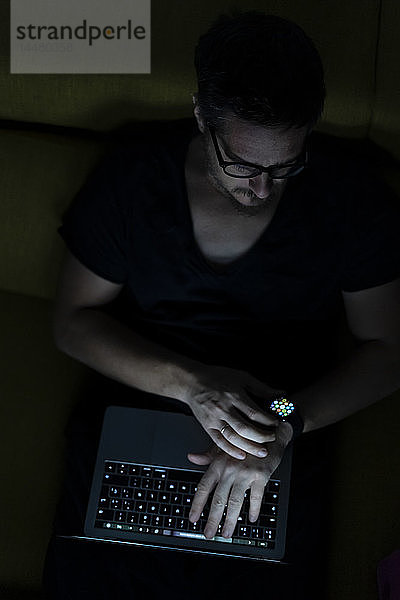 Mann sitzt nachts mit Laptop und Smartwatch auf dem Sofa