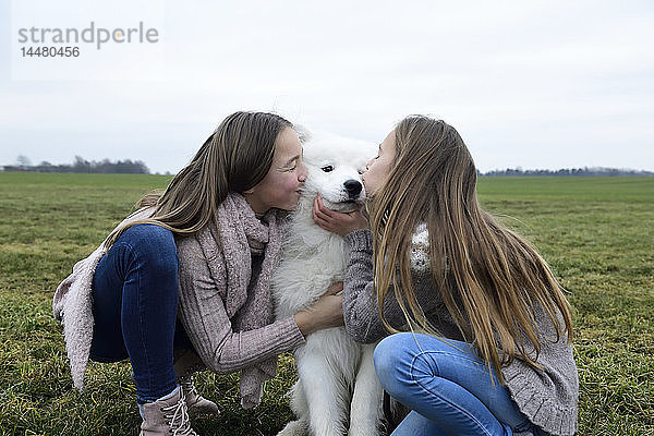 Zwei Mädchen kauern auf einer Wiese und küssen einen Hund