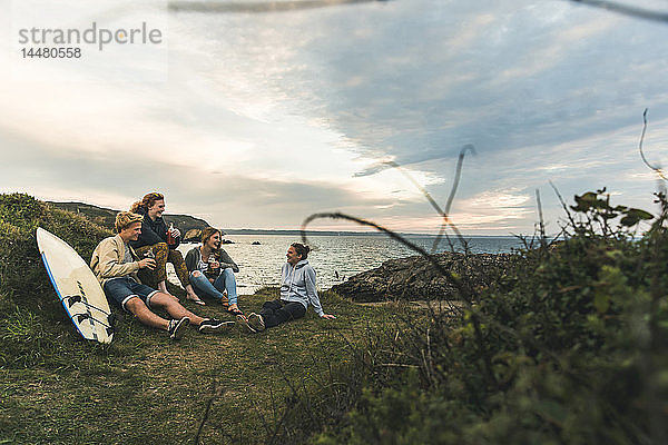 Glückliche Freunde mit Surfbrett und Getränken beim geselligen Beisammensein an der Küste bei Sonnenuntergang