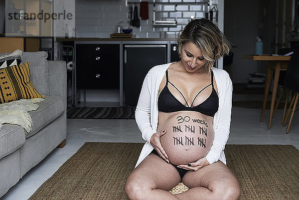 Schwangere Frau sitzt zu Hause auf dem Boden und schaut auf Strichmarken auf ihrem Bauch