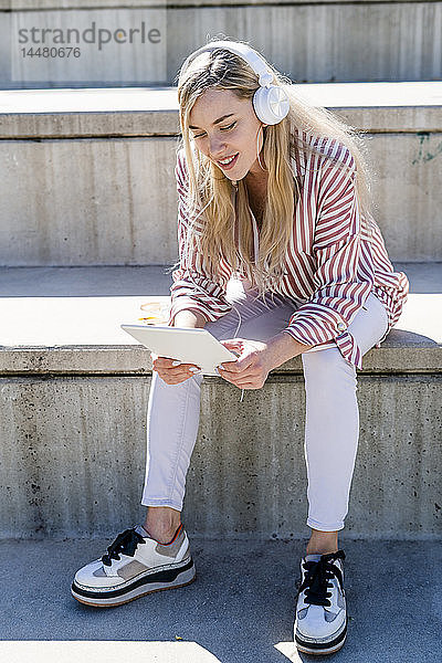 Porträt einer blonden jungen Frau  die auf einer Treppe im Freien sitzt und ein digitales Tablett und Kopfhörer benutzt