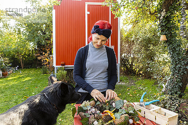 Lächelnde ältere Frau mit rot gefärbtem Haar mit Hund bastelt im Garten an der Herbstdekoration