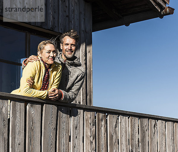 Auf dem Balkon einer Berghütte lehnendes Ehepaar  die Arme um den Kopf gelegt