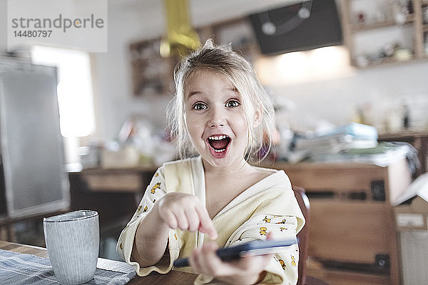 Porträt eines aufgeregten kleinen Mädchens in der Küche  das auf ein Smartphone zeigt