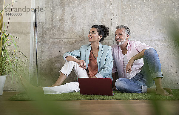 Gelegenheits-Geschäftsmann und -Geschäftsfrau sitzen auf Kunstrasen in einem Loft und teilen sich einen Laptop