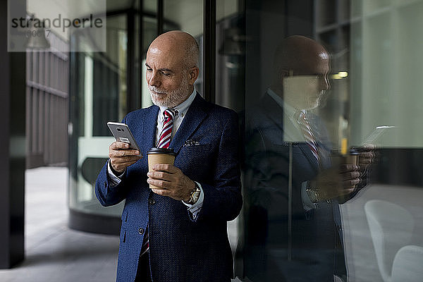 Leitender Geschäftsmann mit Handy und Kaffee zum Mitnehmen an der Glasfront