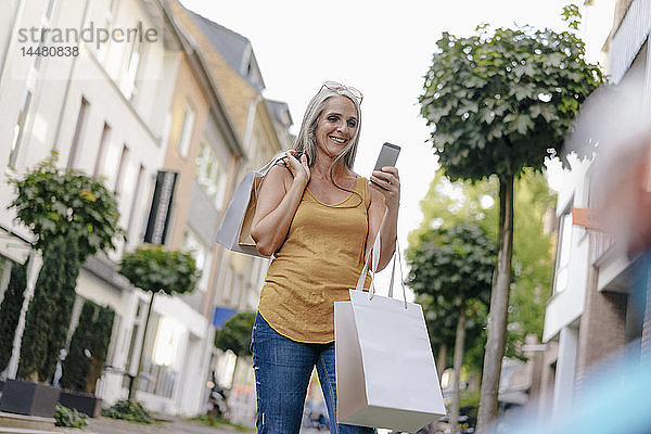 Lächelnde Frau auf der Straße  die Einkaufstüten trägt und auf ihr Handy schaut