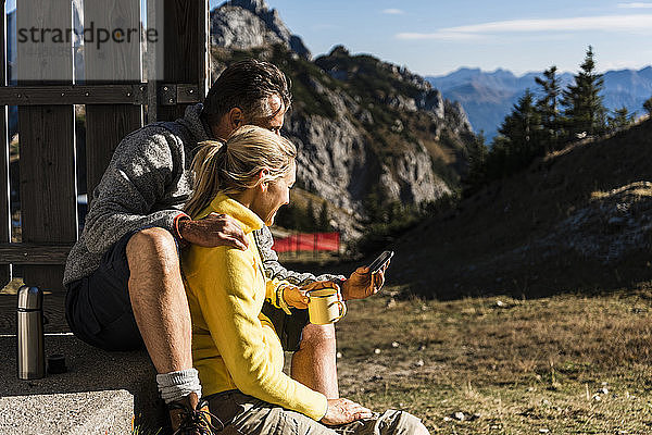 Wanderndes Ehepaar sitzt vor Berghütte  macht Pause  schaut auf Smartphone