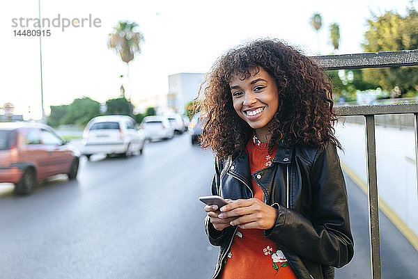 Porträt einer lächelnden jungen Frau mit Smartphone am Straßenrand stehend