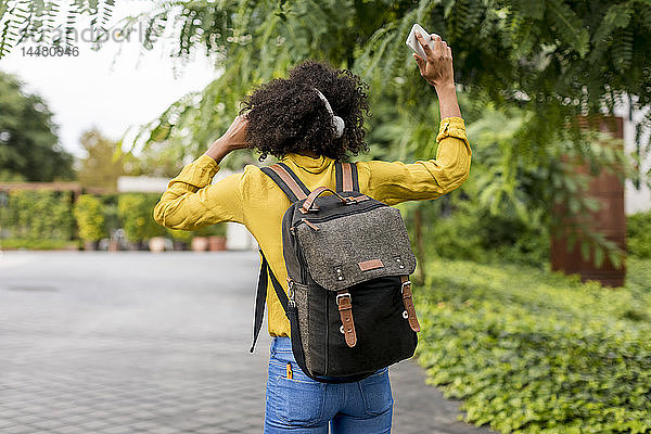 Rückenansicht einer Frau mit Rucksack  die im Freien mit Kopfhörern und Smartphone Musik hört