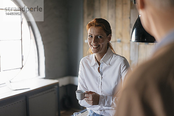 Lächelnde Geschäftsfrau mit einer Tasse Kaffee im Büro  die einen älteren Mann ansieht
