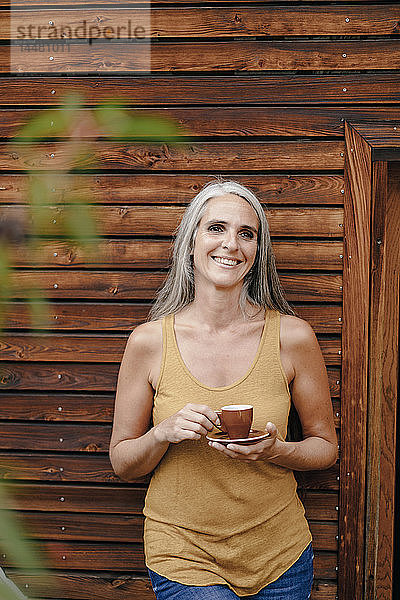 Porträt einer glücklichen reifen Frau mit Kaffeetasse vor einer Holzfassade