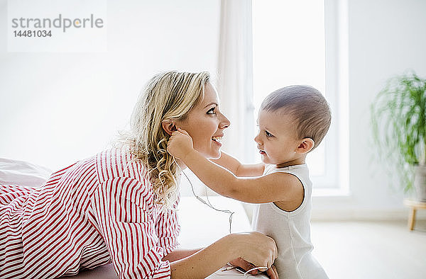 Lächelnde Mutter und Kleinkind-Sohn benutzen Kopfhörer im Schlafzimmer zu Hause