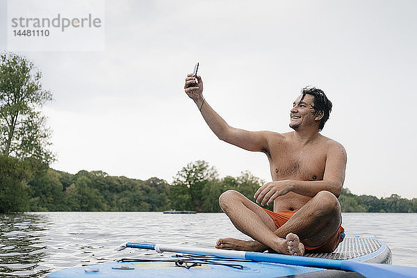 Mann sitzt auf einem SUP-Brett auf einem See und macht ein Selfie