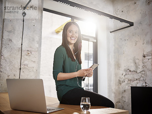 Lächelnde Frau sitzt im Büro auf dem Schreibtisch und hört Musik