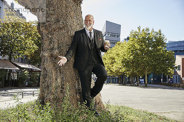 Eleganter Geschäftsmann lehnt sich in der Stadt an einen Baum und trinkt Kaffee