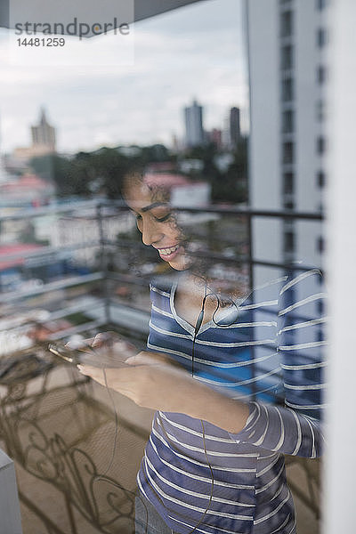Lächelnde junge Frau hinter Fensterscheibe mit Smartphone und Kopfhörern