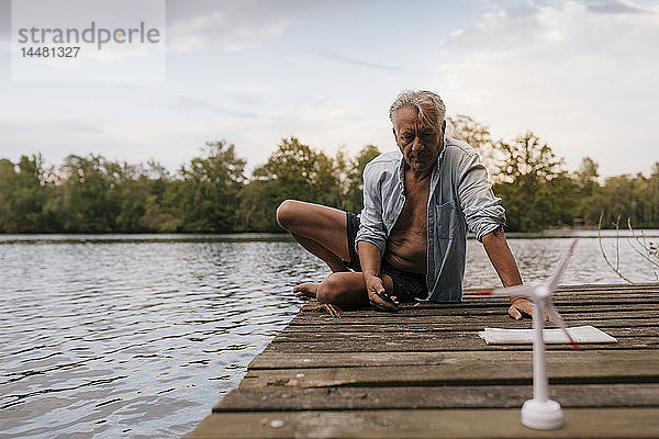 Älterer Mann sitzt auf einem Steg an einem See mit einem Modell einer kleinen Windkraftanlage