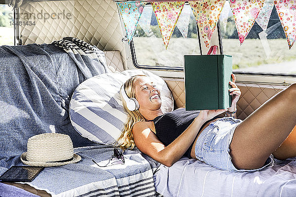 Glückliche Frau mit Kopfhörern liegt in einem Lieferwagen und liest ein Buch