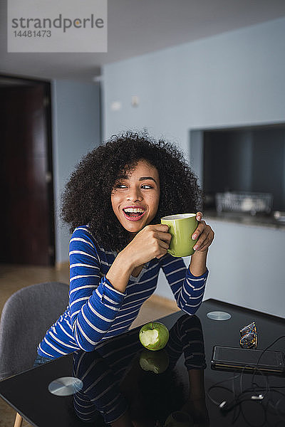 Porträt einer lachenden jungen Frau  die zu Hause Kaffee trinkt und einen Apfel isst