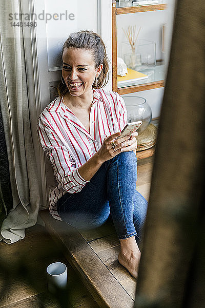Lachende Frau sitzt zu Hause mit Handy auf dem Boden