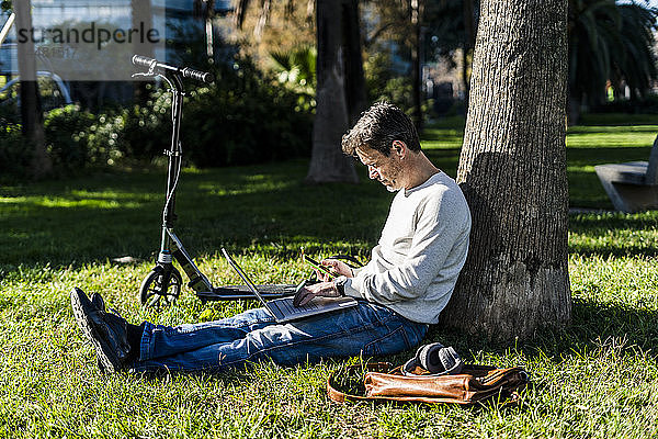zufälliger Geschäftsmann  der auf einem Rasen in einem Park sitzt und einen Laptop benutzt