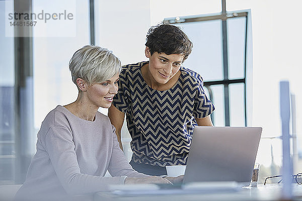 Zwei lächelnde Geschäftsfrauen teilen sich den Laptop am Schreibtisch im Büro