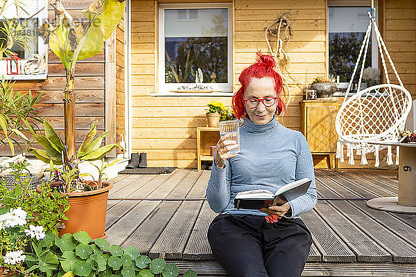 Porträt einer lächelnden älteren Frau mit rot gefärbten Haaren  die auf der Terrasse vor ihrem Haus sitzt und ein Buch liest