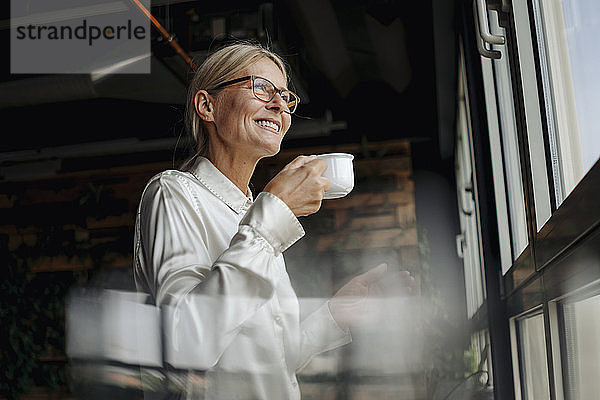 Lächelnde Geschäftsfrau mit einer Tasse Kaffee  die aus dem Fenster schaut