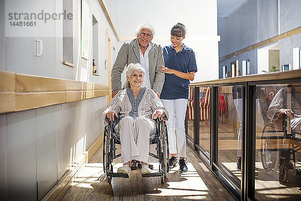Gereatrische Krankenschwester  die älteren Männern und Frauen hilft  den Korridor hinunterzugehen