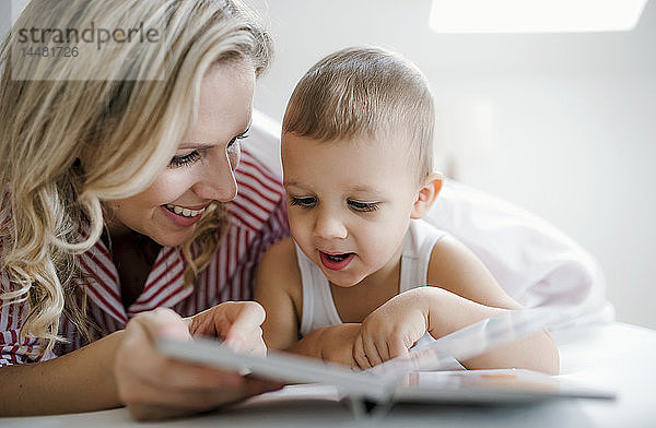 Lächelnde Mutter und Kleinkind-Sohn liegen zu Hause im Bett und lesen ein Buch