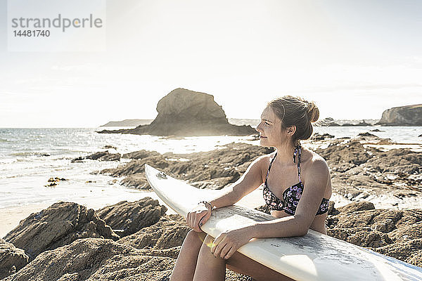 Junge Frau mit Surfbrett entspannt am Strand  auf einem Felsen sitzend