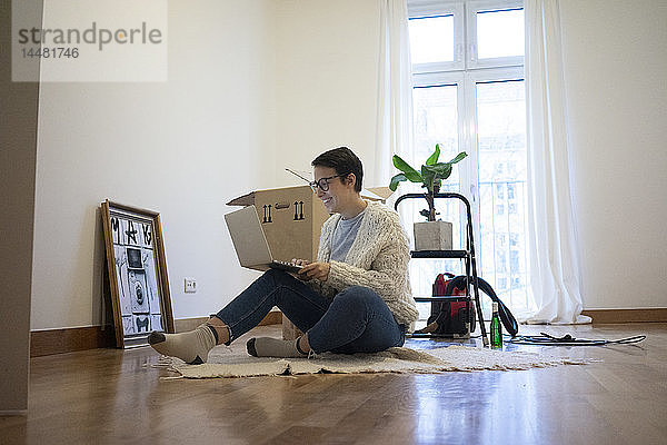 Junge Frau sitzt auf dem Boden ihres neuen Hauses und benutzt einen Laptop