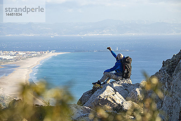 Spanien  Andalusien  Tarifa  Mann auf einer Wanderung an der Küste  der auf einem Felsen sitzt und sich ein Selfie