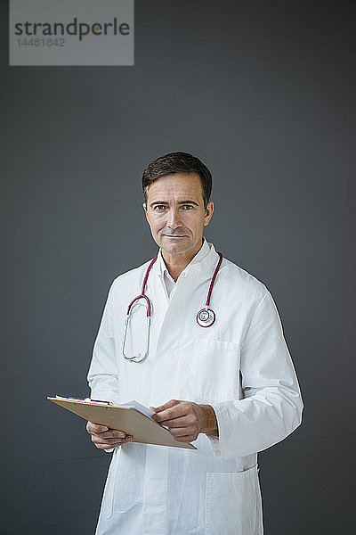 Porträt eines selbstbewussten Arztes  der ein Klemmbrett an einer grauen Wand hält
