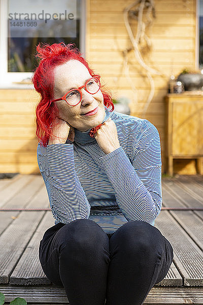 Porträt einer lächelnden älteren Frau mit rot gefärbten Haaren  die auf der Terrasse vor ihrem Haus sitzt und sich entspannt