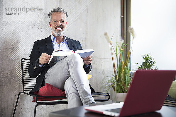 Porträt eines glücklichen Geschäftsmannes  der an einer Betonwand sitzt und ein Buch liest
